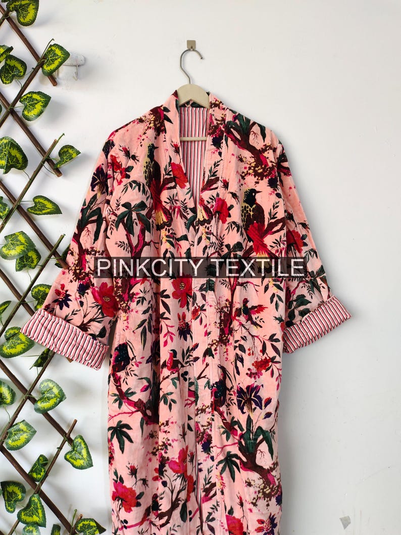 May include: A pink floral robe with a belt, hanging on a wooden hanger. The robe features a bird and floral pattern in red, green, and black. The sleeves are cuffed with a striped fabric. The text "PINKCITY TEXTILE" is visible.