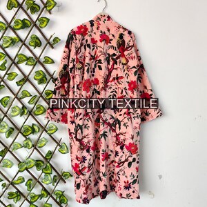 May include: A light pink robe with a floral and bird pattern. The robe is hanging on a silver hanger against a white wall. The floral pattern includes red flowers and green leaves. The text "PINKCITY TEXTILE" is visible at the bottom.