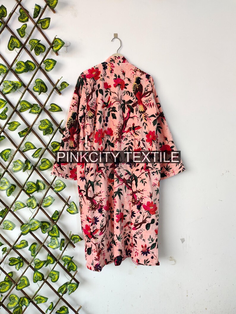 May include: A light pink robe with a floral and bird pattern. The robe is hanging on a silver hanger against a white wall. The floral pattern includes red flowers and green leaves. The text "PINKCITY TEXTILE" is visible at the bottom.