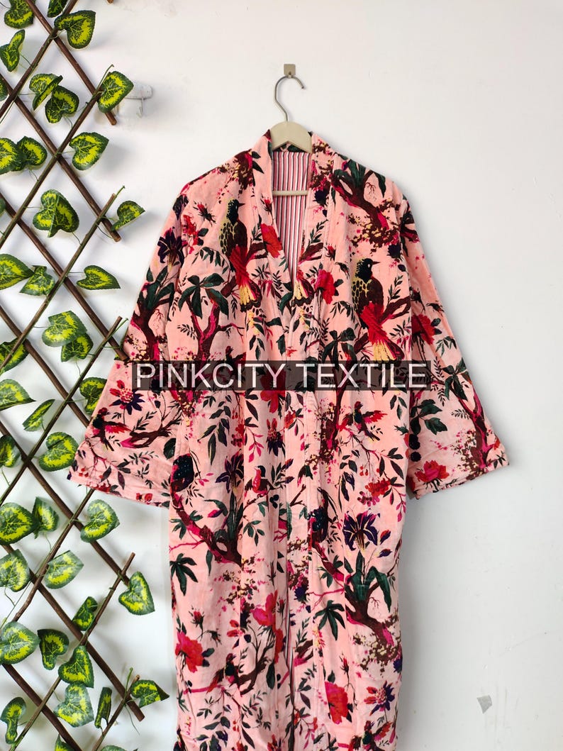 May include: A pink floral print robe with a bird and floral pattern. The robe has long sleeves and is hanging on a wooden hanger. The text "PINKCITY TEXTILE" is visible on the robe.