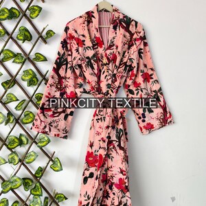 May include: A pink floral print robe with a belt, hanging on a hanger. The robe features a vibrant pattern of red flowers and green leaves against a light pink background. The text "PINK CITY TEXTILE" is printed on the robe.