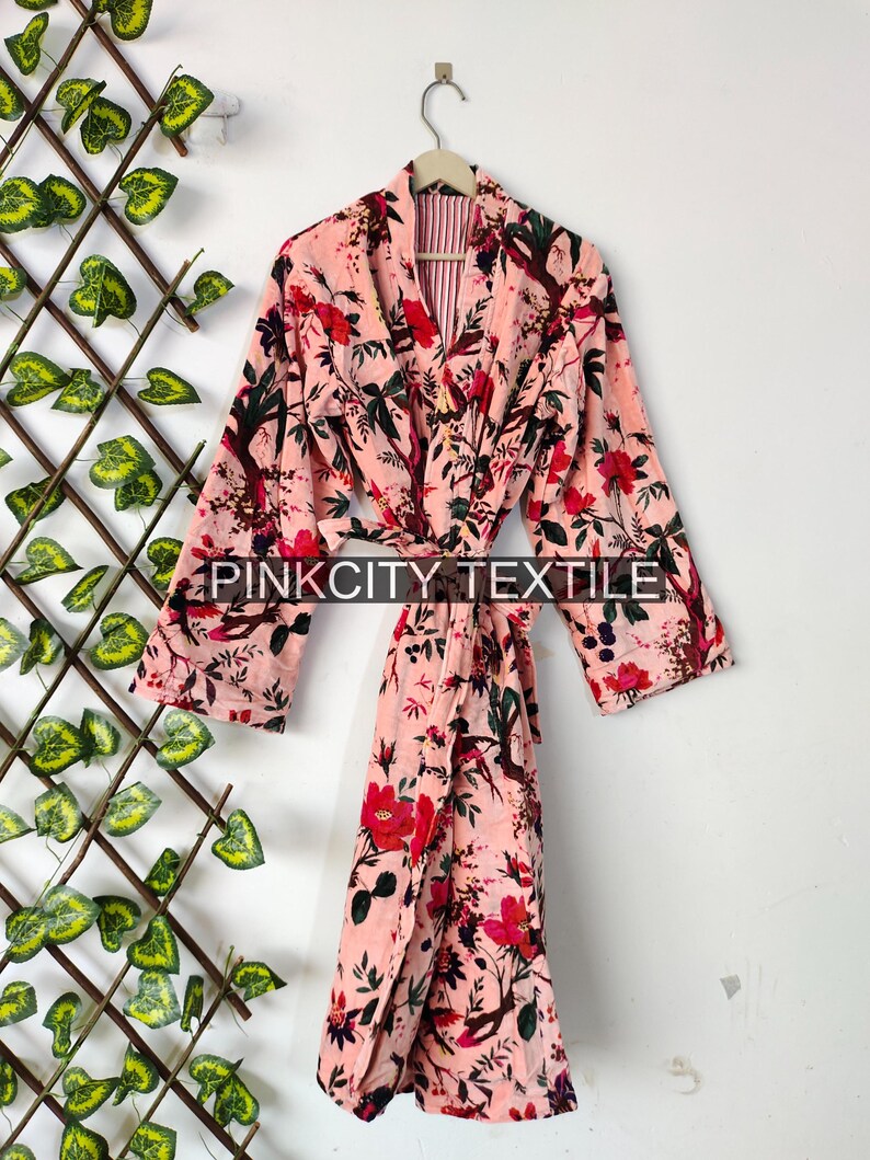 May include: A pink floral print robe with a belt, hanging on a hanger. The robe features a vibrant pattern of red flowers and green leaves against a light pink background. The text "PINK CITY TEXTILE" is printed on the robe.