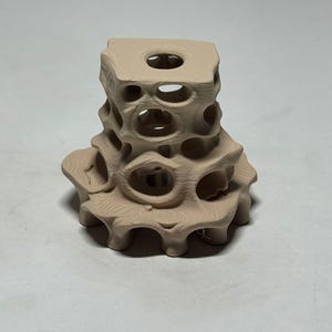 May include: A beige, abstract sculpture with a porous, geometric design. The sculpture has a square top with a central hole and a rounded base. The sides feature numerous circular openings, creating a textured, openwork appearance. The piece is likely made of ceramic or a similar material.