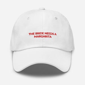 May include: White baseball cap with the red text "THE BRIDE NEEDS A MARGARITA". The cap has a curved brim and a six-panel design. A fun accessory for a bachelorette party or bridal event.