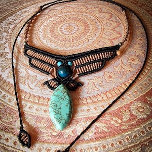 Può includere: Una collana macramè fatta a mano con un grande pendente in pietra turchese. La collana ha un design intrecciato nero e beige con accenti di perline blu e di legno. La collana è esposta su uno sfondo di tessuto fantasia.
