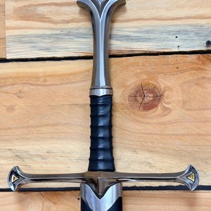 Pode incluir: Uma espada com pomo e guarda de metal prateado polido com detalhes triangulares dourados. O cabo é revestido com material preto texturizado e a lâmina está inserida numa bainha preta com uma ponteira de metal prateado.