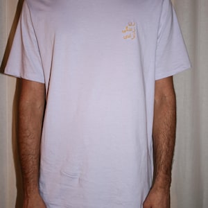 Puede incluir: Una camiseta de manga corta de color lavanda claro con cuello redondo y un peque&ntilde;o texto bordado en amarillo en escritura persa en la parte superior izquierda del pecho. La prenda es de tela lisa y tiene un corte est&aacute;ndar y relajado.