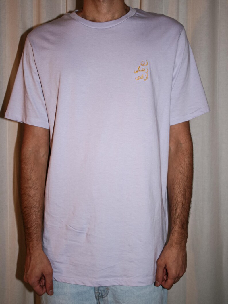Puede incluir: Una camiseta de manga corta de color lavanda claro con cuello redondo y un peque&ntilde;o texto bordado en amarillo en escritura persa en la parte superior izquierda del pecho. La prenda es de tela lisa y tiene un corte est&aacute;ndar y relajado.