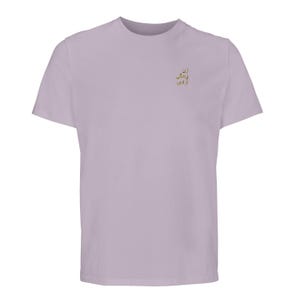 Puede incluir: Una camiseta de manga corta en color lavanda claro con un peque&ntilde;o bordado dorado en la parte superior izquierda del pecho. El bordado consta de tres l&iacute;neas de texto en persa, que se lee como: Zan, Zendegi, Azadi.