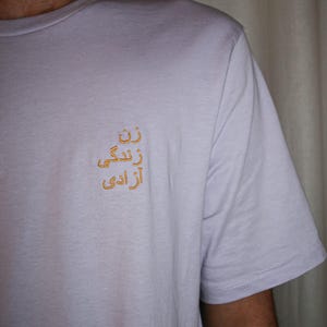 Puede incluir: Una camiseta de manga corta de color lavanda claro con un bordado dorado en el pecho izquierdo. El bordado consta de tres l&iacute;neas de escritura persa, dispuestas verticalmente, que se leen como "Zan, Zendegi, Azadi".