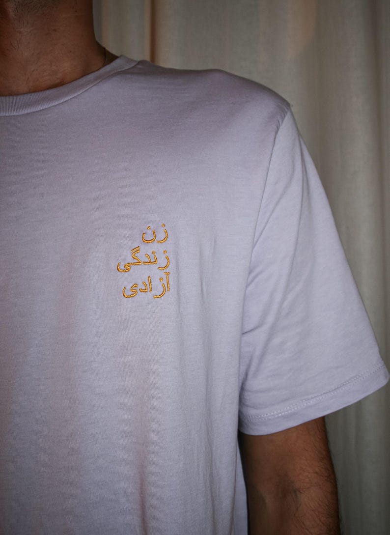 Puede incluir: Una camiseta de manga corta de color lavanda claro con un bordado dorado en el pecho izquierdo. El bordado consta de tres l&iacute;neas de escritura persa, dispuestas verticalmente, que se leen como "Zan, Zendegi, Azadi".