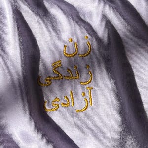 Puede incluir: Una tela de color lavanda claro con texto bordado en hilo dorado en escritura persa. El bordado consta de tres l&iacute;neas de texto apiladas verticalmente que dicen "زن زندگی آزادی" en un estilo sans-serif. La tela presenta pliegues y arrugas.