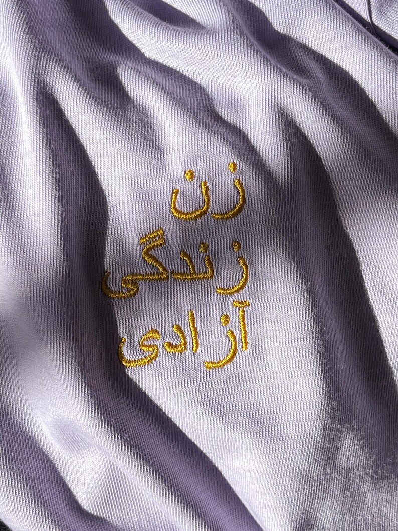 Puede incluir: Una tela de color lavanda claro con texto bordado en hilo dorado en escritura persa. El bordado consta de tres l&iacute;neas de texto apiladas verticalmente que dicen "زن زندگی آزادی" en un estilo sans-serif. La tela presenta pliegues y arrugas.