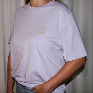 Puede incluir: Una camiseta de manga corta en color lavanda claro con un peque&ntilde;o bordado dorado en la parte superior izquierda del pecho. El bordado consta de tres l&iacute;neas de escritura persa, que se lee como 'Zan, Zendegi, Azadi' en una fuente sencilla.