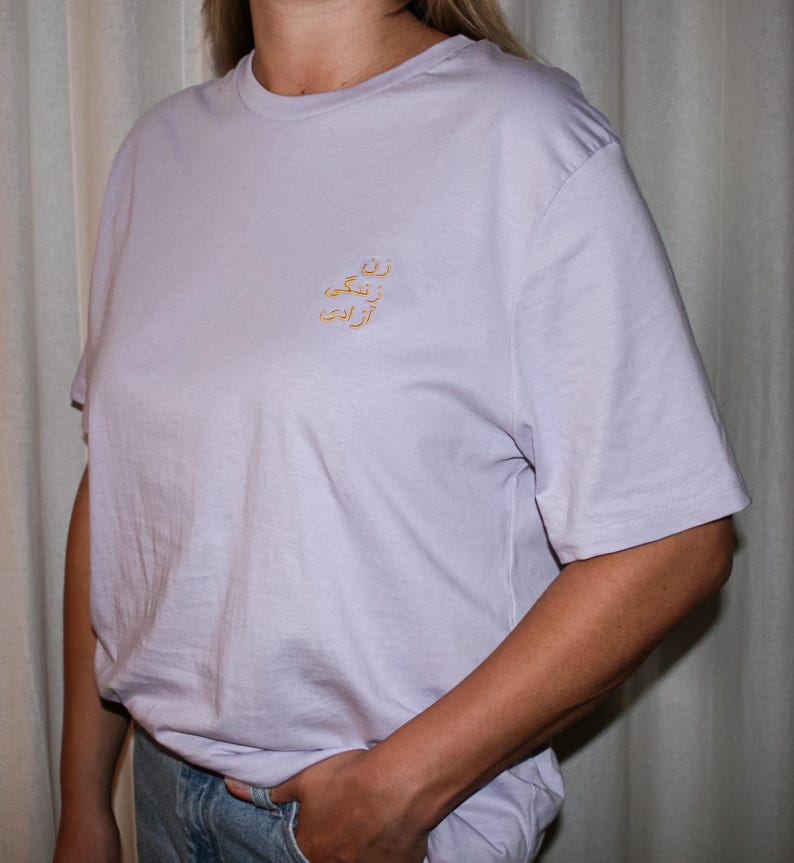 Puede incluir: Una camiseta de manga corta en color lavanda claro con un peque&ntilde;o bordado dorado en la parte superior izquierda del pecho. El bordado consta de tres l&iacute;neas de escritura persa, que se lee como 'Zan, Zendegi, Azadi' en una fuente sencilla.