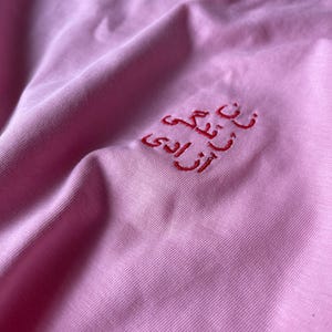 Puede incluir: Una camiseta de color rosa claro con texto bordado en rojo en escritura persa. El bordado est&aacute; situado en la zona superior del pecho de la prenda. El tejido tiene una textura lisa y las costuras son claramente visibles sobre el fondo rosa.