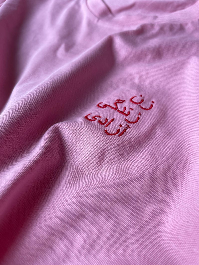 Puede incluir: Una camiseta de color rosa claro con texto bordado en rojo en escritura persa. El bordado est&aacute; situado en la zona superior del pecho de la prenda. El tejido tiene una textura lisa y las costuras son claramente visibles sobre el fondo rosa.