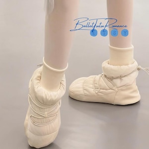 May include: Cream-colored ballet bootie slippers with quilted texture and lace-up detail. The slippers are worn with off-white tights. The text "Ballet Tutu Romance" is in the upper center of the image.