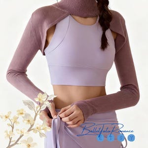 May include: A lilac-colored sports bra and matching skirt are paired with a mauve-colored long-sleeved shrug. The shrug has a high neck and cut-out shoulder design. The image has the text "Ballet Tutu Romance" in the bottom right corner.