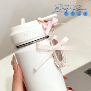 Puede incluir: Una botella de agua blanca con un llavero dorado que sostiene un dije de zapatilla de ballet en miniatura con una cinta rosa. La botella tiene una tapa blanca y una banda plateada. El texto "BalletTutuRomance" está en la esquina superior derecha.