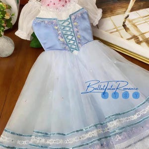 Puede incluir: Un vestido de tutú de ballet azul claro con un corpiño de satén y una falda de tul en capas. El corpiño presenta un diseño con cordones y bordados florales. El vestido tiene mangas cortas y abullonadas y está adornado con lentejuelas. El texto "Ballet Tutu Romance" es visible.
