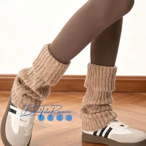 Op de afbeelding: Tan gebreide beenwarmers gedragen over bruine leggings en witte sneakers met zwarte strepen. De beenwarmers zijn geribbeld en hebben een gevouwen manchet. De sneakers hebben een grijze neus en een bruine zool.