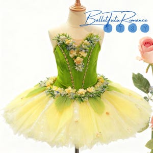 以下が含まれることがあります： 緑と黄色のバレエチュチュ、花の装飾付き。 ボディスは鮮やかな緑色で、繊細な花とビーズのアクセントで飾られています。 チュチュスカートは黄色から白に変わり、重ね着効果を生み出しています。 「Ballet Tutu Romance」の文字が見えます。