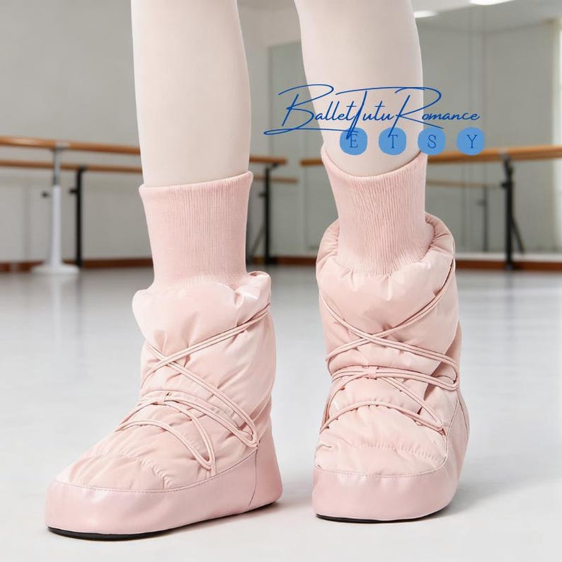 May include: A pair of light pink quilted ballet boots with criss-cross laces. The boots have a soft, padded appearance and are worn with matching pink leg warmers. The background shows a ballet studio with a barre.