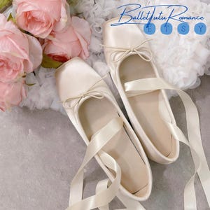 May include: Two pairs of ivory satin ballet flats with ribbon ties and small bows. The shoes are displayed on a light grey surface, with a backdrop of pink roses and white tulle. The text "BalletTutu Romance" is visible in the upper right corner.