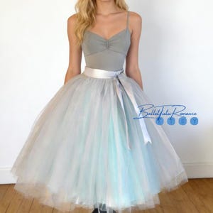 Może przedstawiać: Szary trykot z ramiączkami spaghetti i dekoltem w kształcie serca w połączeniu z warstwową spódnicą z tiulu w odcieniach szarości i błękitu. Biały satynowy pasek z kokardą podkreśla talię. Widoczny jest tekst "BalletTutu Romance".