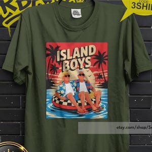 May include: Olive green t-shirt featuring a graphic of two people on an inflatable ring with the text "ISLAND BOYS" against a sunset backdrop. The shirt also has the text "SUB URBAN" in yellow graffiti style.