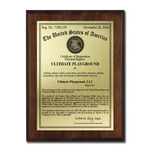 May include: A framed certificate from the United States of America, featuring a gold-colored plate with black text. The certificate is for "ULTIMATE PLAYGROUND" and lists clothing items. The frame is a dark brown color.