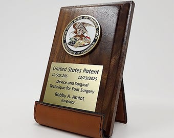 May include: A wooden plaque with a gold-colored seal and a brass plate. The seal reads "United States Patent and Trademark Office." The brass plate has text that reads "United States Patent" and other details. A brown leather support is at the bottom.