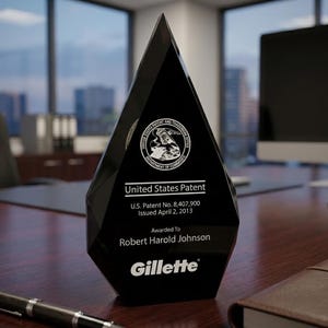 以下が含まれることがあります： 幾何学的なデザインの黒いクリスタルアワード。アワードには「United States Patent」と「Gillette」の文字が記載されています。米国特許第8,407,900号は2013年4月2日にRobert Harold Johnsonに発行されました。アワードは木製の机の上にあります。