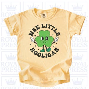 May include: A light yellow t-shirt featuring a cartoon shamrock character with the text "WEE LITTLE HOOLIGAN" in a circular design. The shamrock has a smiling face, peace signs, and star accents.