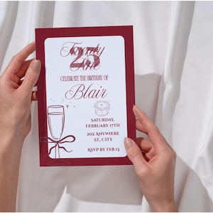 May include: A burgundy birthday invitation with the text "Twenty Fine" and "Celebrate the Birthday of Blair." The invitation includes a champagne glass illustration and event details. The invitation is held against a white fabric background.