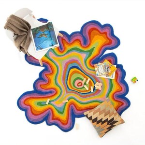 Op de afbeelding: Een kleurrijk, abstract tapijt met een gelaagd ontwerp in tinten blauw, paars, roze, geel en groen. Een witte stoel met een bruine deken, een kussen met patroon, boeken en houten blokken op een witte achtergrond.