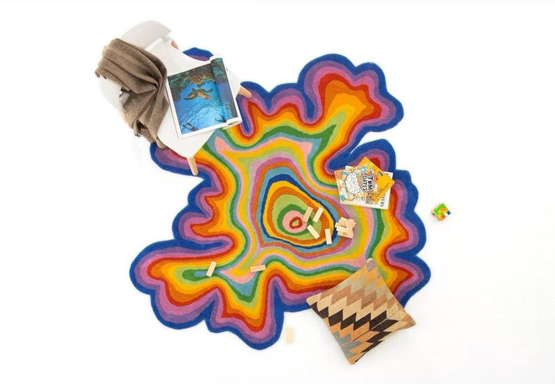 Op de afbeelding: Een kleurrijk, abstract tapijt met een gelaagd ontwerp in tinten blauw, paars, roze, geel en groen. Een witte stoel met een bruine deken, een kussen met patroon, boeken en houten blokken op een witte achtergrond.