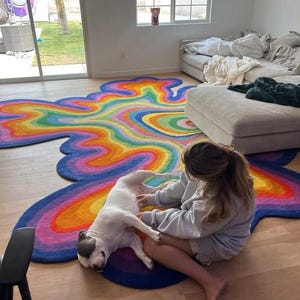 Op de afbeelding: Een kleurrijk, abstract tapijt met een regenboogontwerp. Het tapijt heeft een vloeiende, wolkachtige vorm met levendige tinten blauw, paars, oranje, geel en groen. Het tapijt ligt op een houten vloer.