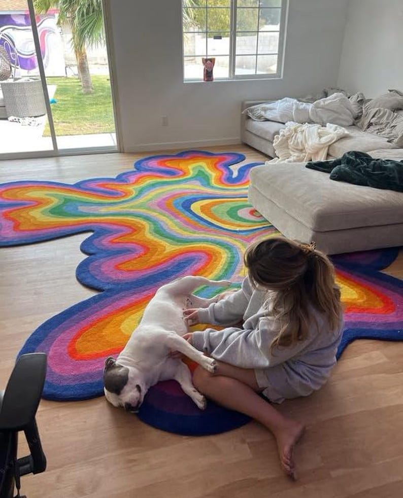Op de afbeelding: Een kleurrijk, abstract tapijt met een regenboogontwerp. Het tapijt heeft een vloeiende, wolkachtige vorm met levendige tinten blauw, paars, oranje, geel en groen. Het tapijt ligt op een houten vloer.