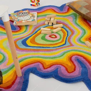 Op de afbeelding: Een kleurrijk, abstract tapijt met een regenboogontwerp. Het tapijt heeft concentrische, golvende lijnen in tinten blauw, paars, roze, geel, oranje en groen. Een houten tafelbeen, een boek en een kussen zijn ook zichtbaar.