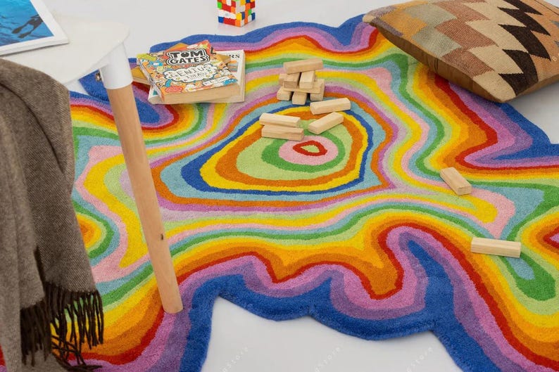 Op de afbeelding: Een kleurrijk, abstract tapijt met een regenboogontwerp. Het tapijt heeft concentrische, golvende lijnen in tinten blauw, paars, roze, geel, oranje en groen. Een houten tafelbeen, een boek en een kussen zijn ook zichtbaar.