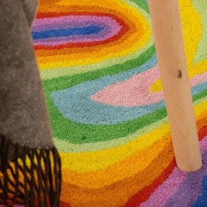 Op de afbeelding: Een kleurrijk, getuft tapijt met een regenboogontwerp. Het tapijt heeft concentrische banden van geel, oranje, rood, roze, groen, blauw en paars. Een houten poot van een tafel of stoel is zichtbaar, samen met een bruine deken met franjes.