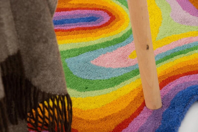 Op de afbeelding: Een kleurrijk, getuft tapijt met een regenboogontwerp. Het tapijt heeft concentrische banden van geel, oranje, rood, roze, groen, blauw en paars. Een houten poot van een tafel of stoel is zichtbaar, samen met een bruine deken met franjes.