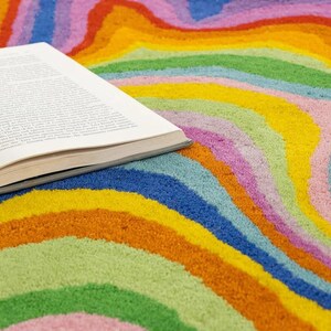 Op de afbeelding: Een open boek ligt op een kleurrijk tapijt met een golvend patroon. Het tapijt heeft een levendig regenboogpatroon met oranje, geel, groen, blauw en roze. De pagina's van het boek zijn zichtbaar.