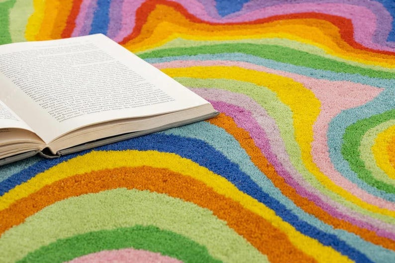 Op de afbeelding: Een open boek ligt op een kleurrijk tapijt met een golvend patroon. Het tapijt heeft een levendig regenboogpatroon met oranje, geel, groen, blauw en roze. De pagina's van het boek zijn zichtbaar.