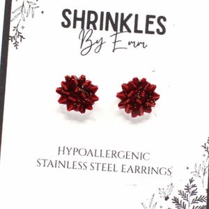 May include: A pair of red bow-shaped stud earrings on a white card. The card has the text "SHRINKLES By Emm" and "HYPOALLERGENIC STAINLESS STEEL EARRINGS". The earrings are a deep red color.