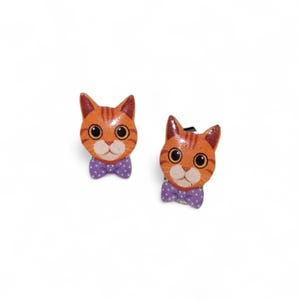 May include: Two orange cat-shaped earrings with black eyes and purple polka dot bow ties. The earrings have a striped pattern and are set against a white background. These cat earrings are a fun accessory.
