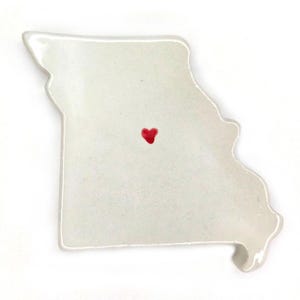 May include: A white ceramic dish in the shape of the state of Missouri, with a red heart marking a location. The dish has a glossy finish and a slightly irregular shape, suggesting it is handmade. The heart is centrally located.