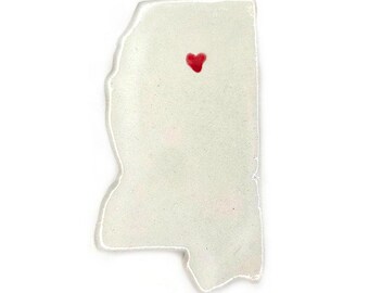 May include: A white, Mississippi-shaped ornament with a small red heart near the top. The ornament has a slightly textured surface and a clear, irregular edge, set against a white background.